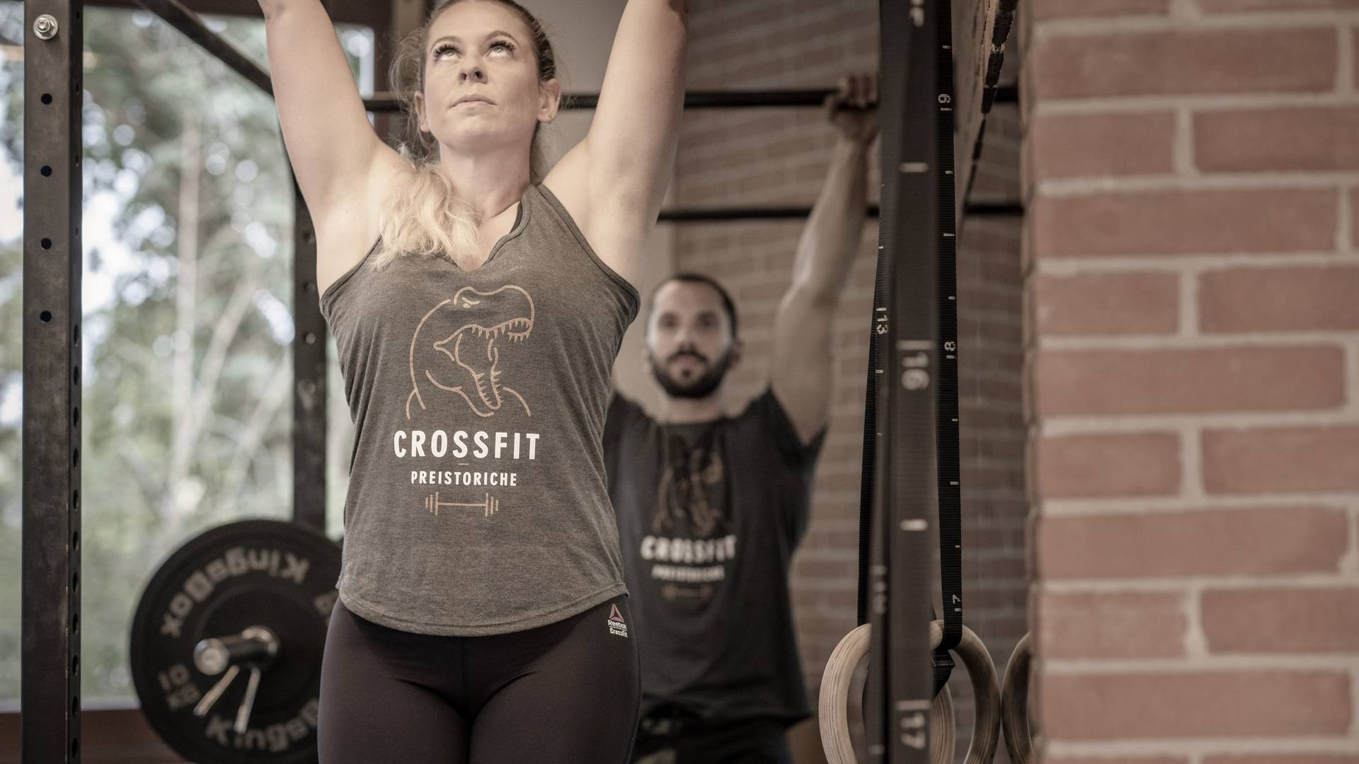 Donna compie un esercizio di trazione alla sbarra in una palestra di CrossFit, con indosso attrezzatura sportiva e concentrazione intensa, su sfondo di una parete in mattoni e attrezzature da allenamento.